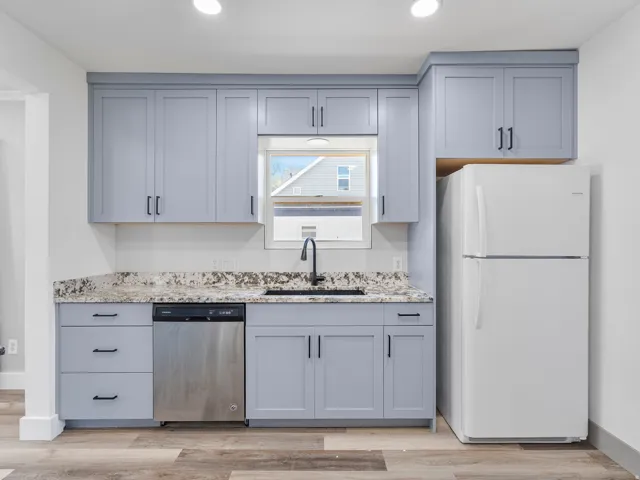 Contemporary kitchen featuring light gray cabinetry with black hardware, stone-finish countertops, stainless steel dishwasher, and wood-finish flooring