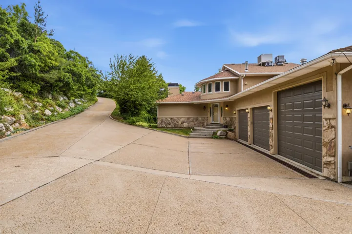 Expansive concrete driveway featuring a three-car garage with individual doors, a stucco exterior with stone accents, and a winding approach surrounded by mature greenery