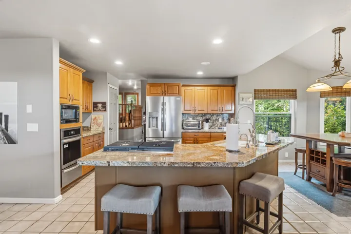 Expansive kitchen featuring a large island with a cooktop, granite countertops, wood cabinetry, stainless steel appliances, and tile flooring