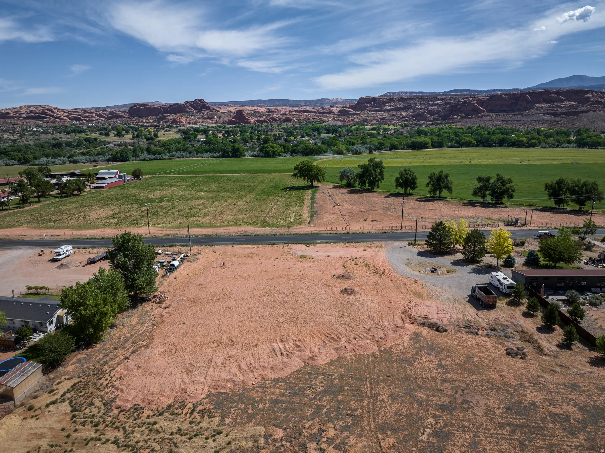 Expansive land parcel featuring a gravel driveway and scattered mature trees, set against a backdrop of distant red rock formations and verdant fields