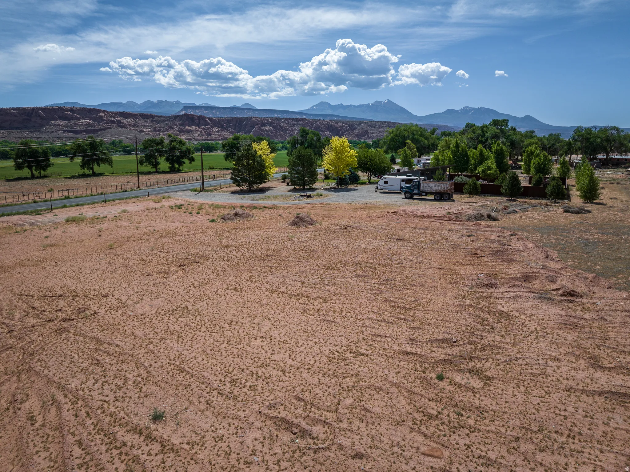 Expansive open land featuring distant mountains, a green valley, mature trees, and a paved road