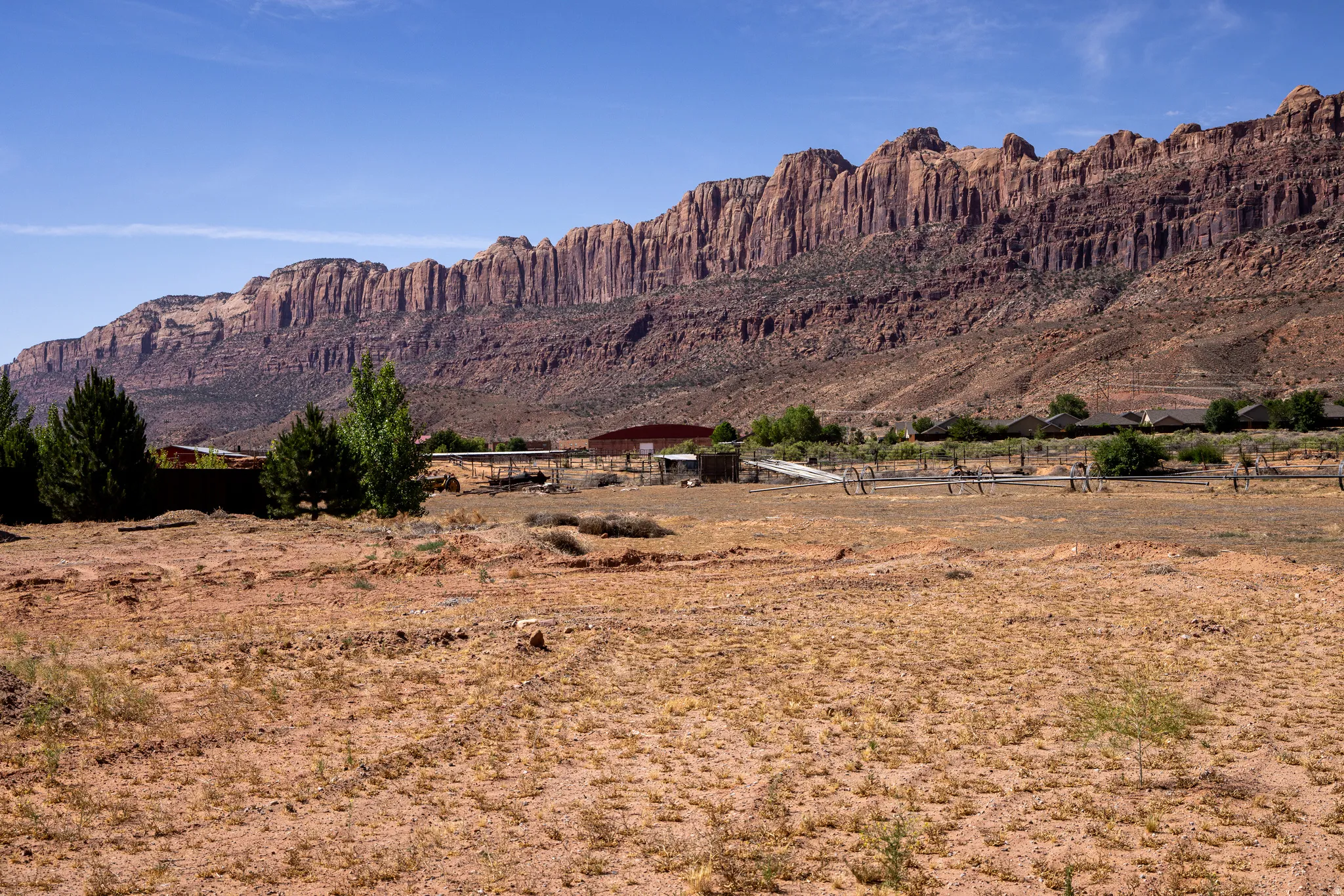 Expansive desert landscape featuring a clear blue sky, distant red rock formations, scattered green trees, and multiple structures