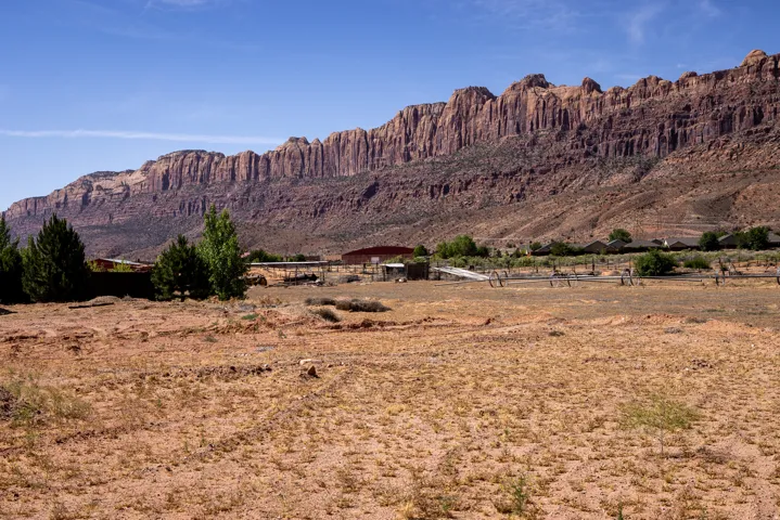 Expansive desert landscape featuring a clear blue sky, distant red rock formations, scattered green trees, and multiple structures