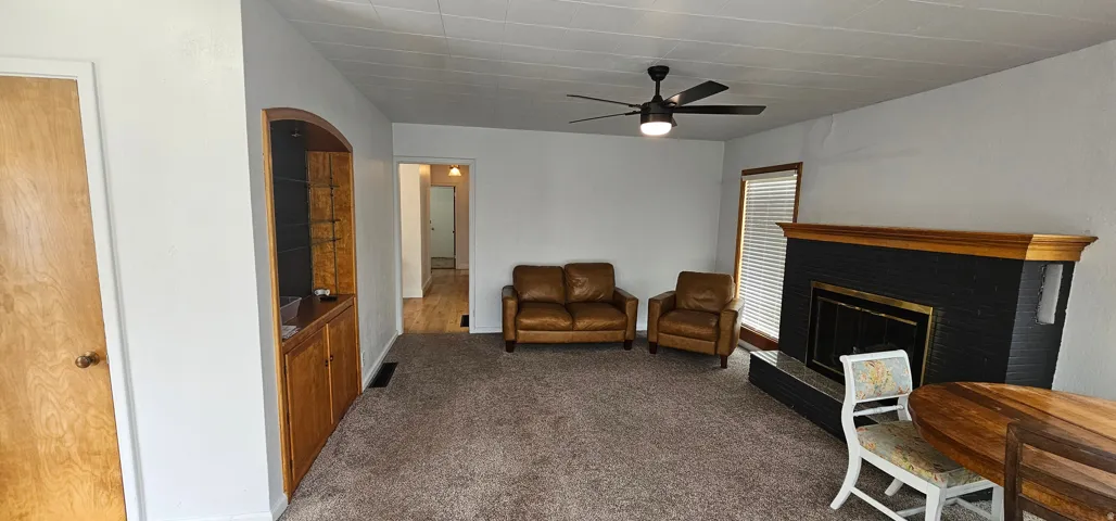 Spacious living area featuring a fireplace with a black surround and wood mantel, neutral carpeting, and a ceiling fan with integrated lighting