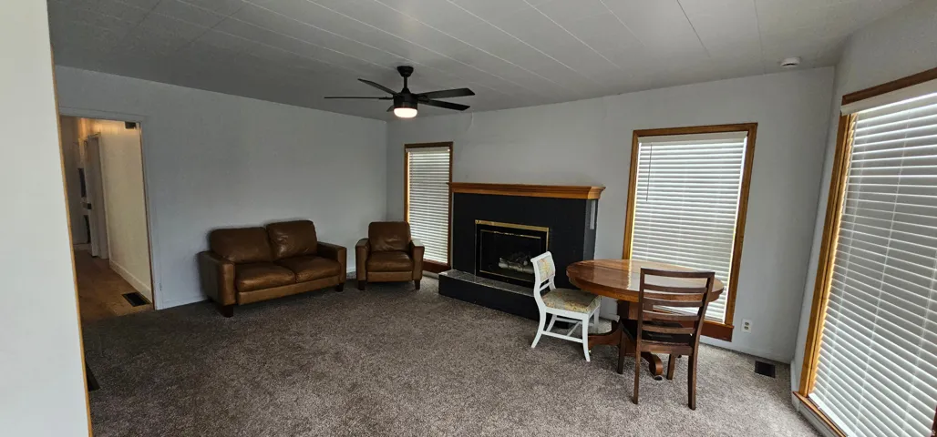 Living area featuring a central fireplace with a dark surround and wood mantel, neutral-toned carpeting, and a ceiling fan with integrated lighting