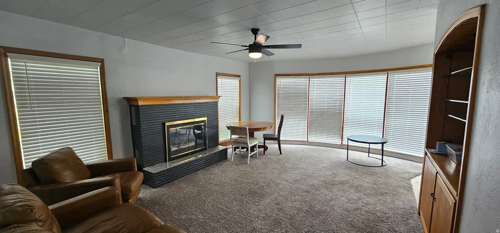 Living area featuring a textured fireplace with a wood mantle, a ceiling fan, and multiple windows with blinds