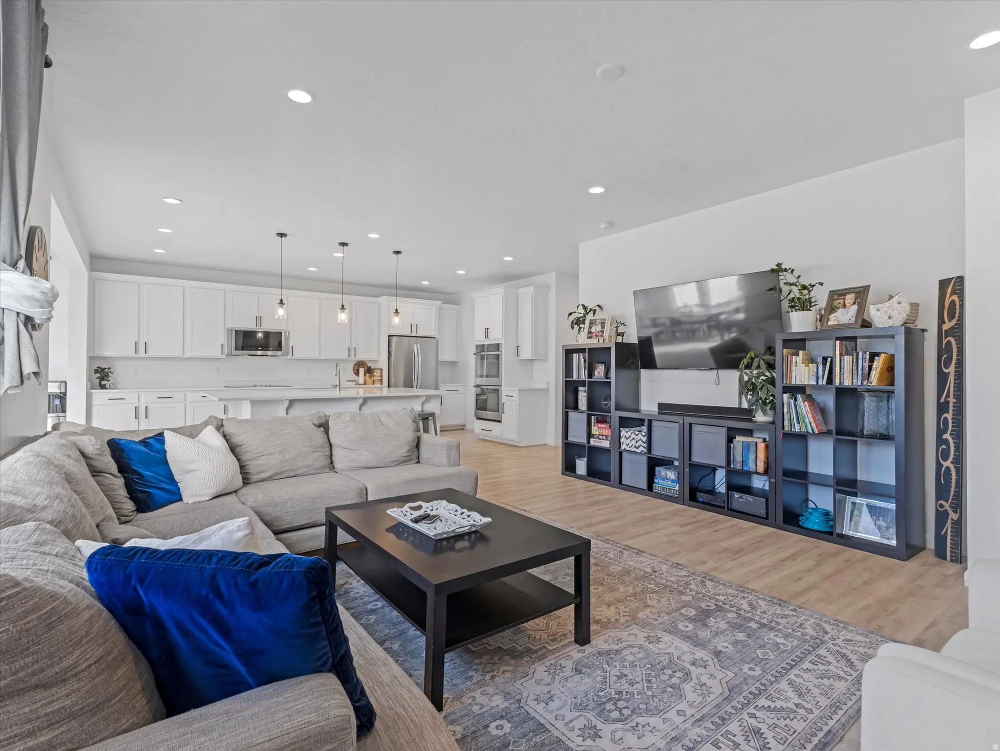 Open-concept living space with light wood-finish flooring, recessed lighting, and a kitchen featuring white cabinetry, a stainless steel refrigerator, and pendant lighting