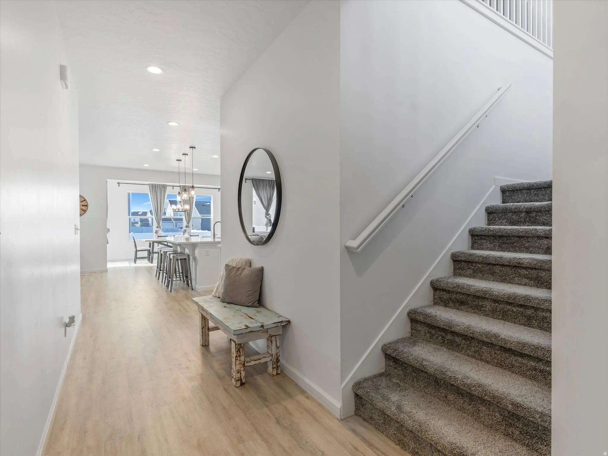 Bright interior hallway featuring wood-finish flooring, a carpeted staircase with a white handrail, recessed lighting, and a sightline to the kitchen