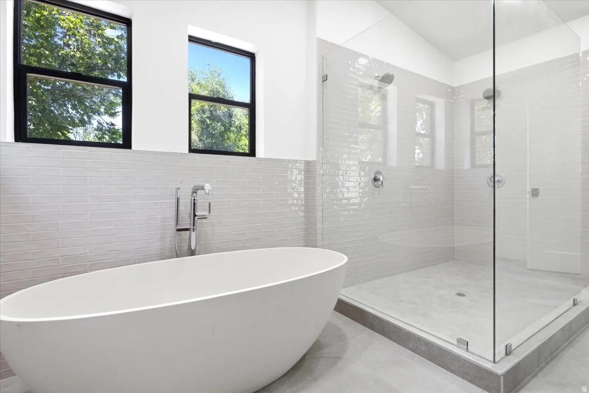 Bathroom featuring a freestanding oval soaking tub with floor-mounted faucet, a spacious glass-enclosed shower with dual showerheads, light-toned horizontal tile work, and two rectangular windows