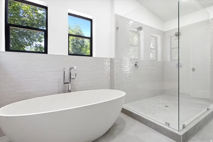 Bathroom featuring a freestanding oval soaking tub with floor-mounted faucet, a spacious glass-enclosed shower with dual showerheads, light-toned horizontal tile work, and two rectangular windows