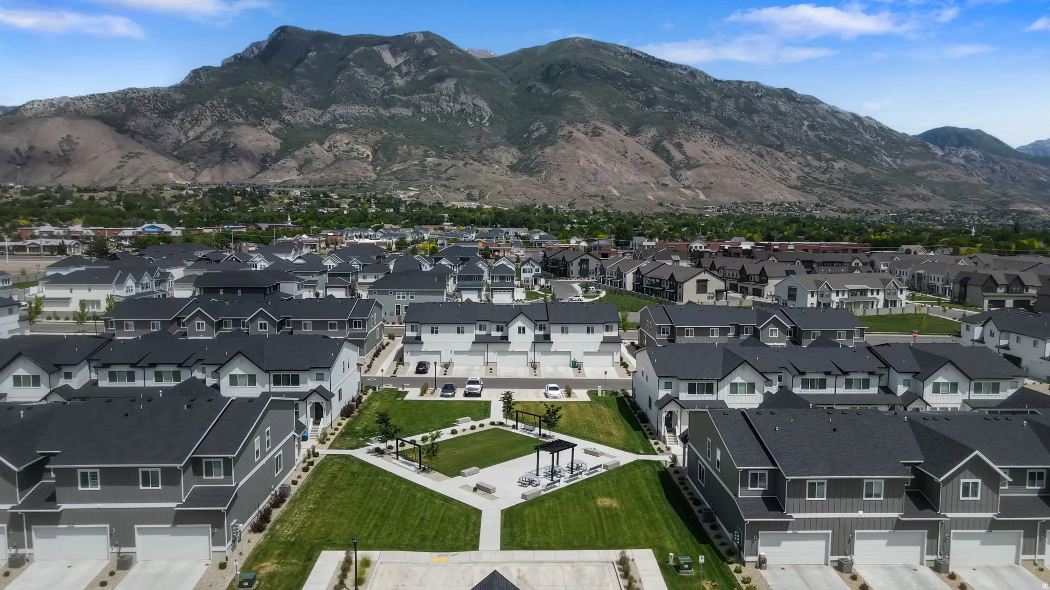 Community green space with concrete pathways and shaded seating areas surrounded by multi-story residences featuring attached garages, dark shingle roofs, and light-colored siding