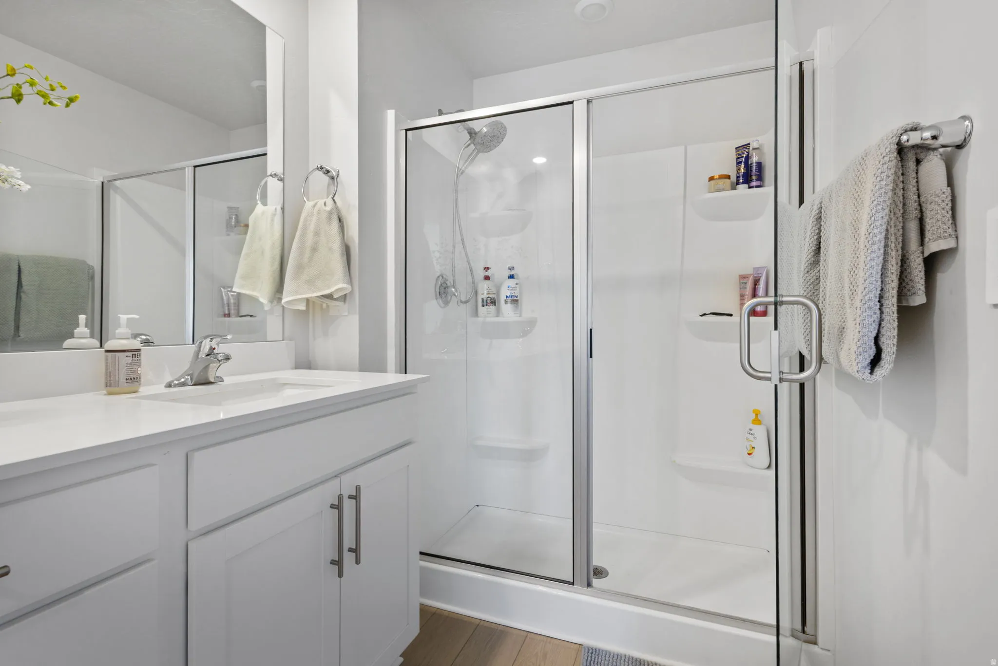 Modern bathroom featuring a glass-enclosed shower with integrated shelving, a sleek single-sink vanity with a light-toned countertop, and wood-finish flooring