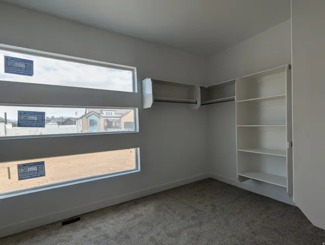 Bedroom featuring three horizontal windows, plush gray carpeting, white baseboards, and a built-in closet system with shelving and hanging rods