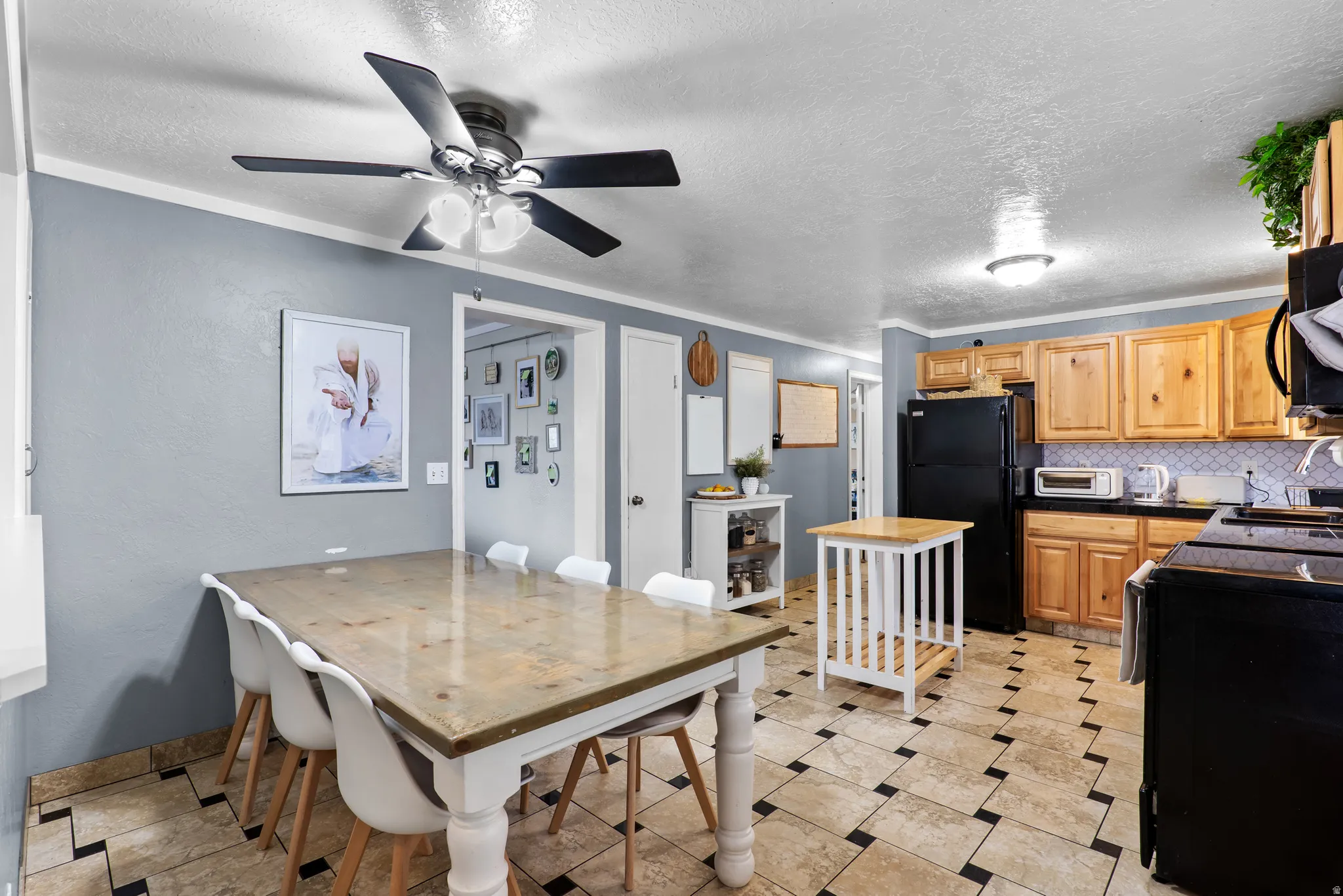 Kitchen and dining area featuring light wood cabinetry, black appliances, a geometric tiled floor, and a ceiling fan with integrated lighting
