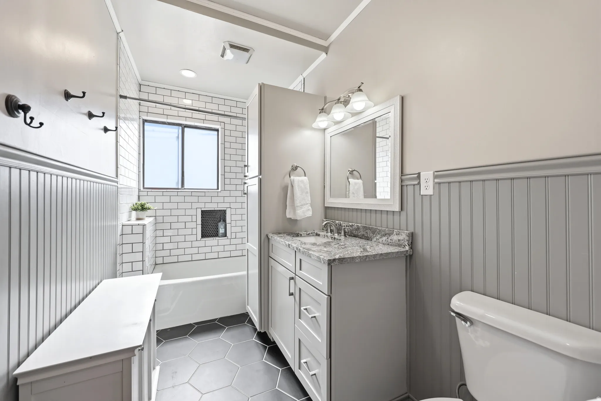 Bathroom featuring a single vanity with a stone countertop, subway tile shower surround with built-in niche, hexagonal floor tiling, wainscoting, and a window