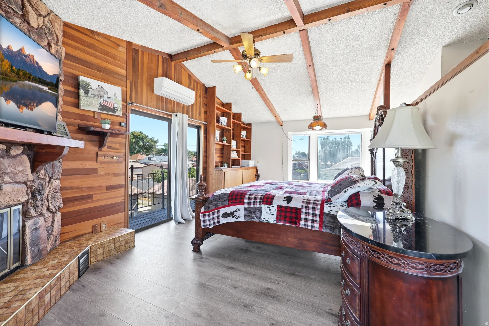 Spacious bedroom with vaulted ceilings, exposed wood beams, wood-finish flooring, a stone-clad fireplace, and exterior access via sliding glass doors