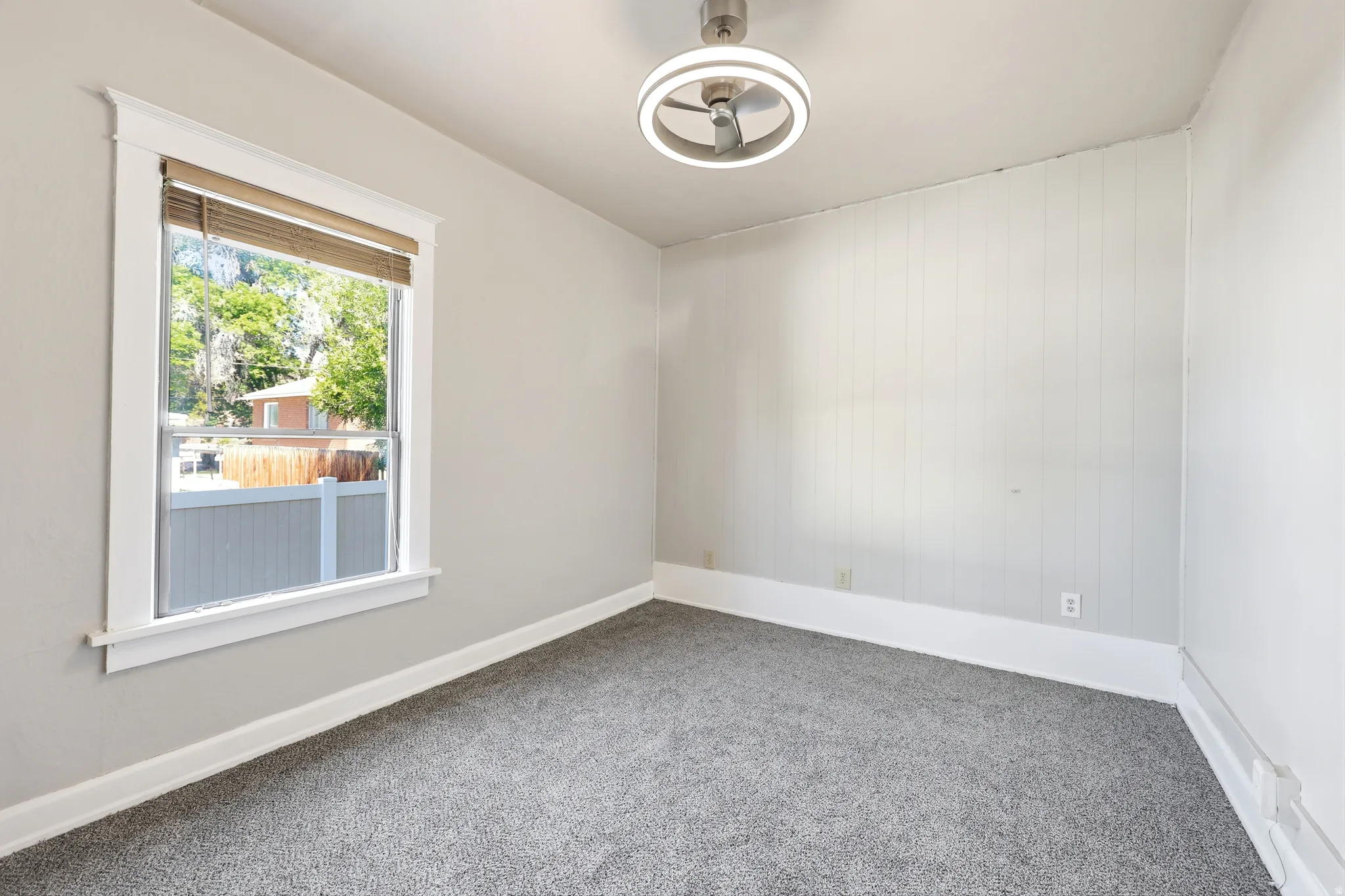 Carpeted interior space featuring a window with white trim, a modern ceiling fan with integrated lighting, and a wall with vertical paneling