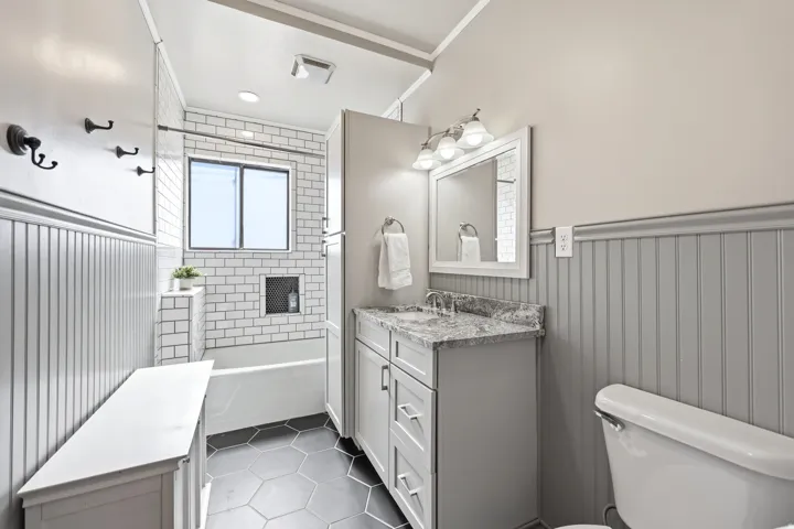 Bathroom featuring a single vanity with a stone countertop, subway tile shower surround with built-in niche, hexagonal floor tiling, wainscoting, and a window