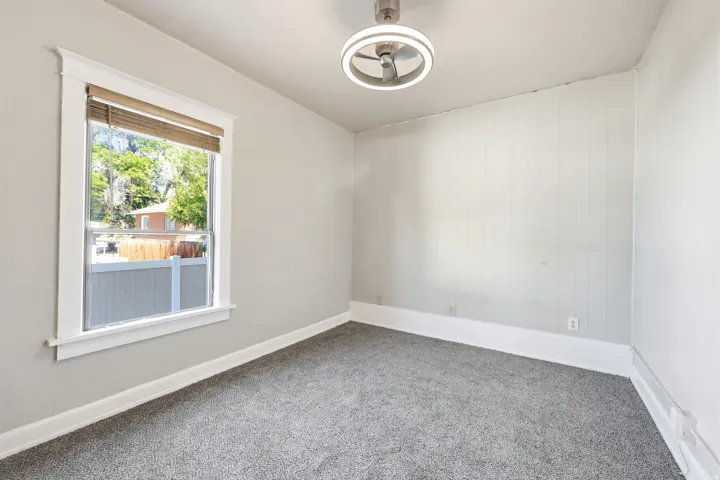 Carpeted interior space featuring a window with white trim, a modern ceiling fan with integrated lighting, and a wall with vertical paneling