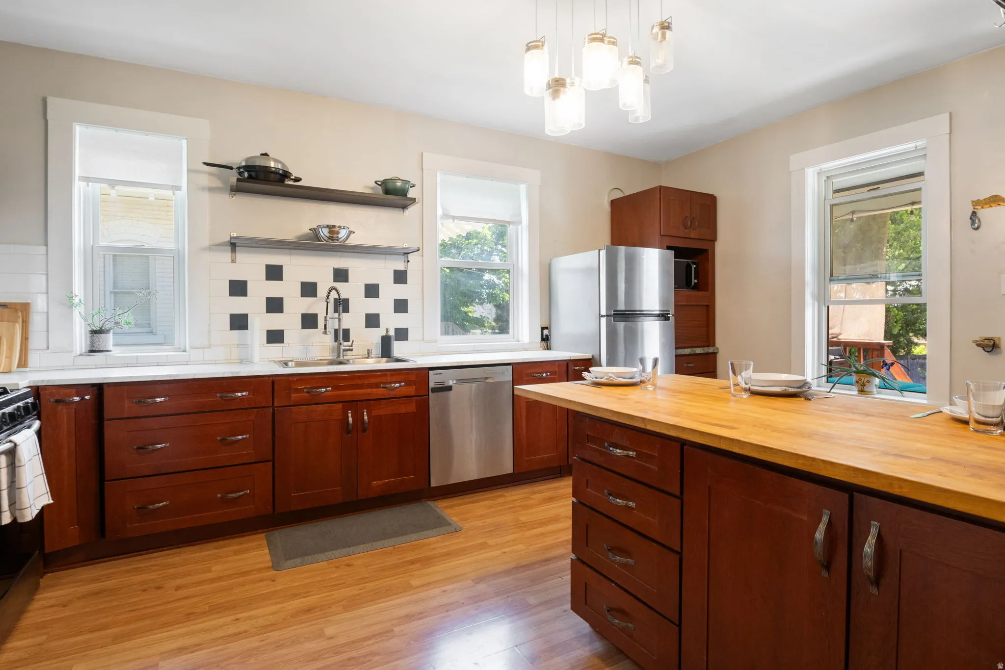 Expansive kitchen featuring a central island with a wood-finish countertop, rich wood-toned cabinetry, stainless steel appliances, and a tiled backsplash with decorative square accents