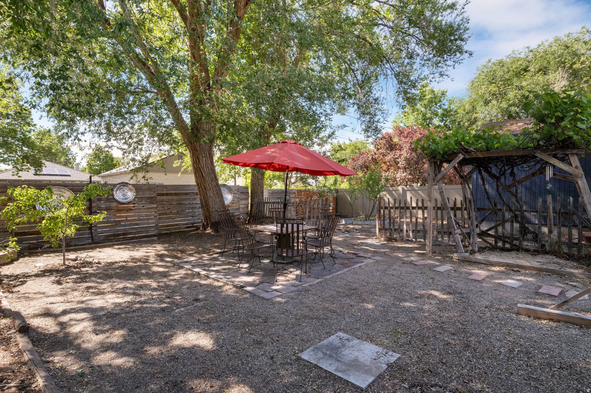 Expansive gravel yard featuring mature trees, a built-in wooden fence, a pergola structure, and stepping stones