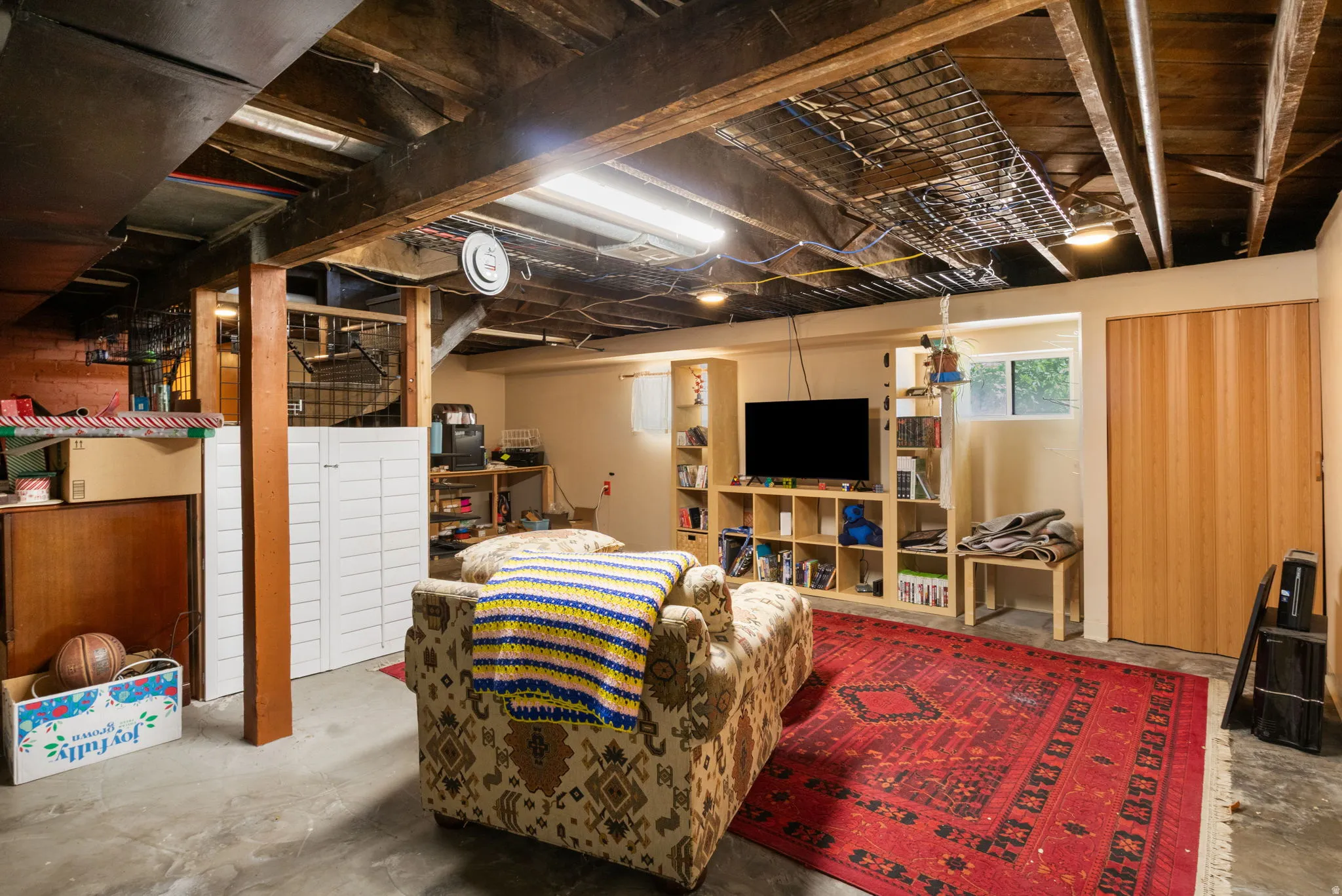 Finished basement area featuring exposed wood beams, concrete flooring, built-in shelving, a natural wood-finish bi-fold door, and a single window