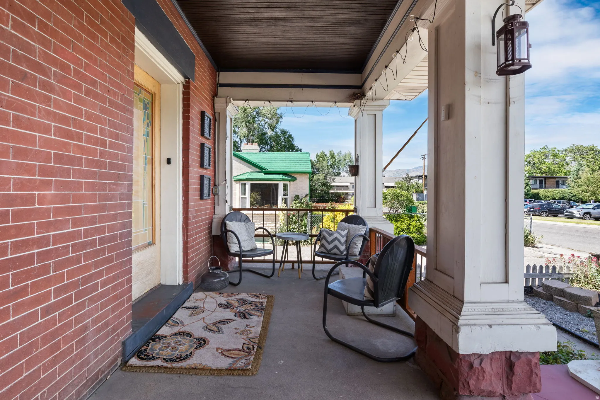 Inviting front porch featuring a brick facade, white architectural columns, a dark wood-finish ceiling, and an outdoor lantern