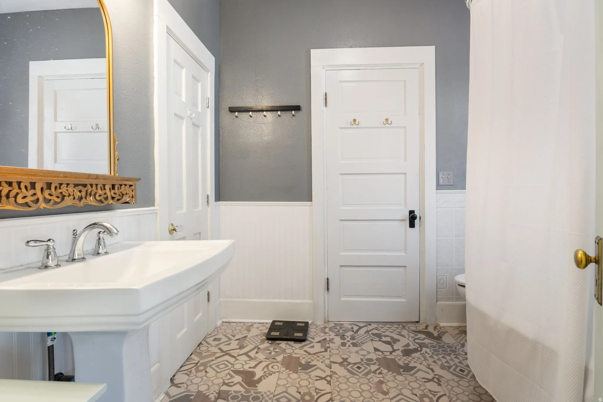 Pedestal sink with chrome fixtures, intricate gold-tone mirror, white beadboard wainscoting, patterned floor tiles, and a white paneled door