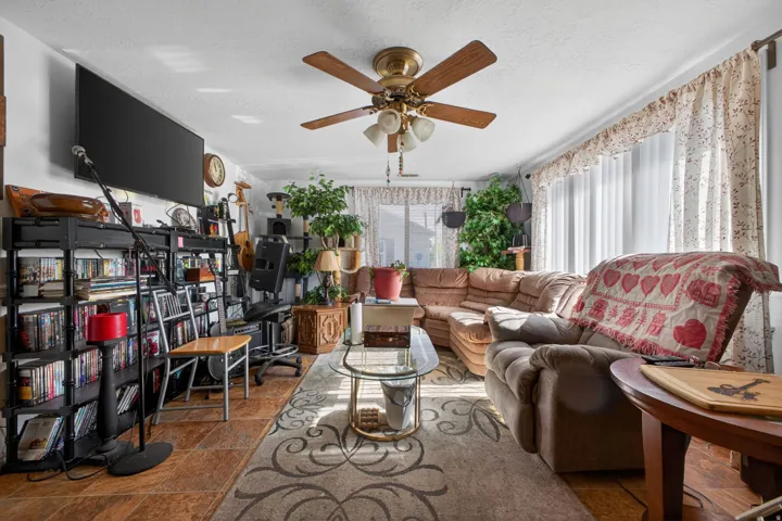 Spacious living area featuring tiled flooring, a ceiling fan with integrated lighting, and multiple windows with vertical blinds