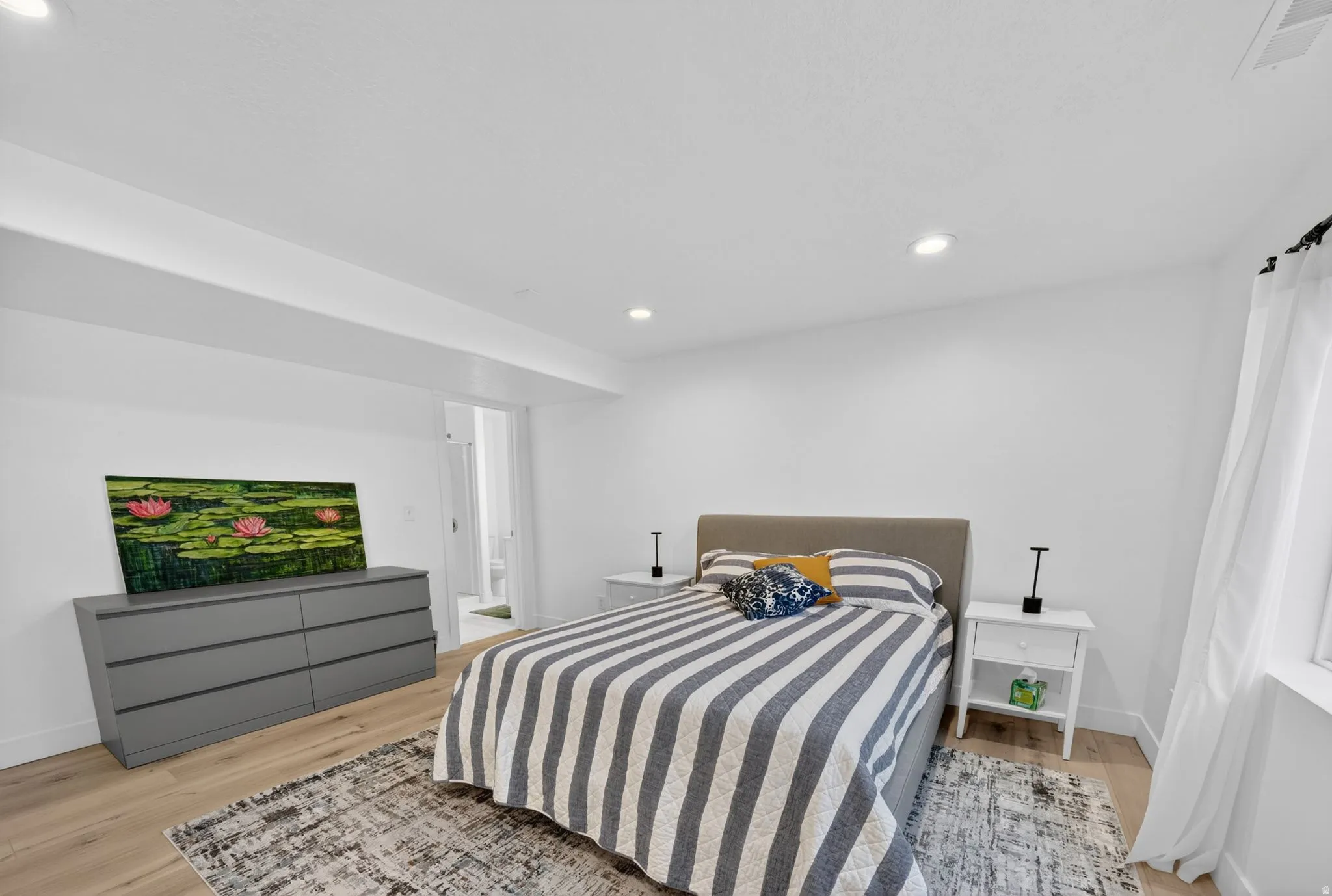 Spacious bedroom featuring light wood-finish flooring, white walls, recessed ceiling lighting, and a window with white drapes