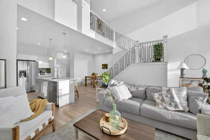 Spacious open-concept living area featuring high ceilings, wood-finish flooring, a modern kitchen with stainless steel appliances and an island, and a prominent staircase with decorative railings