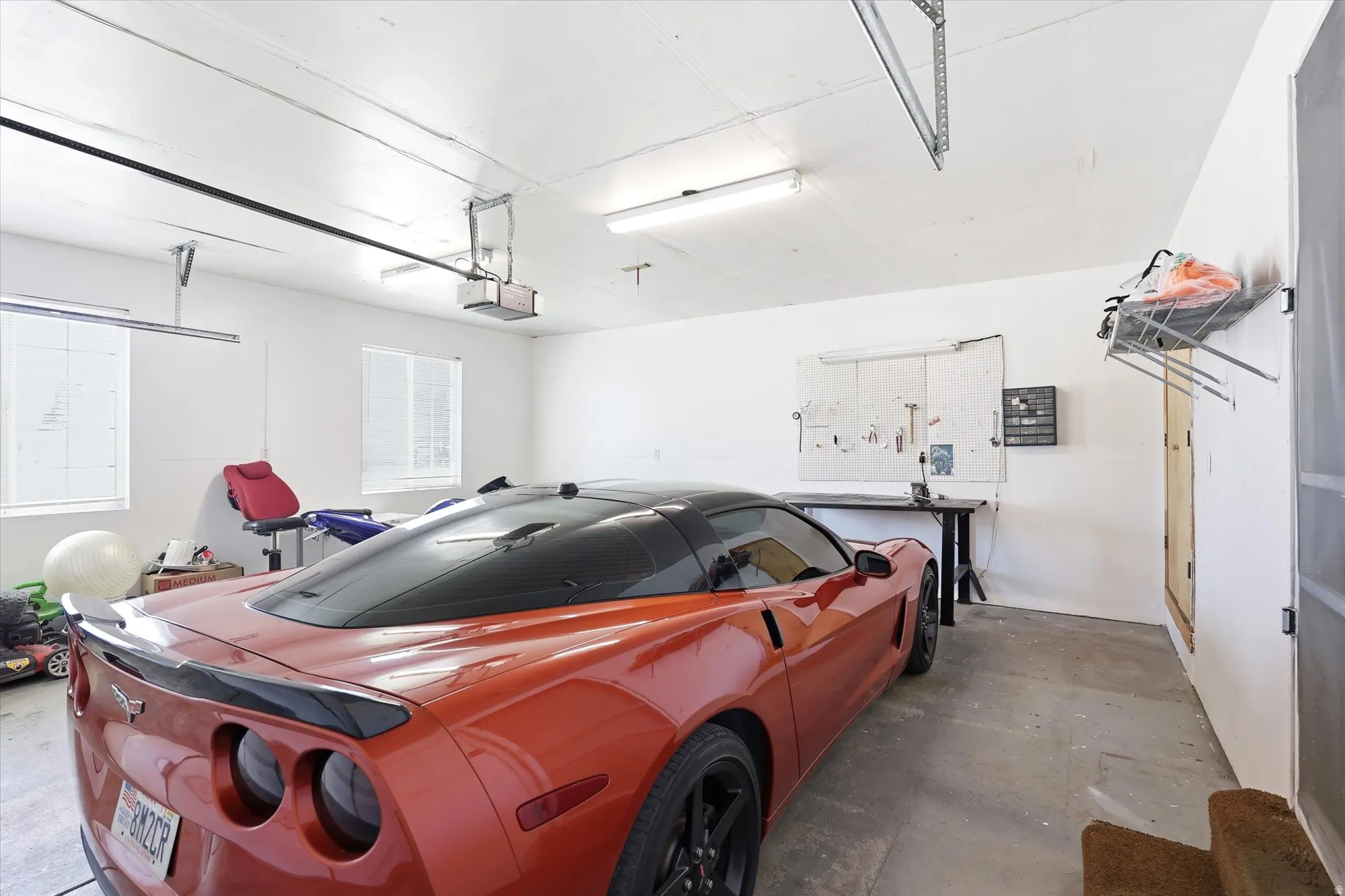 Spacious garage featuring white walls, concrete flooring, two windows with blinds, a built-in workbench, and overhead fluorescent lighting