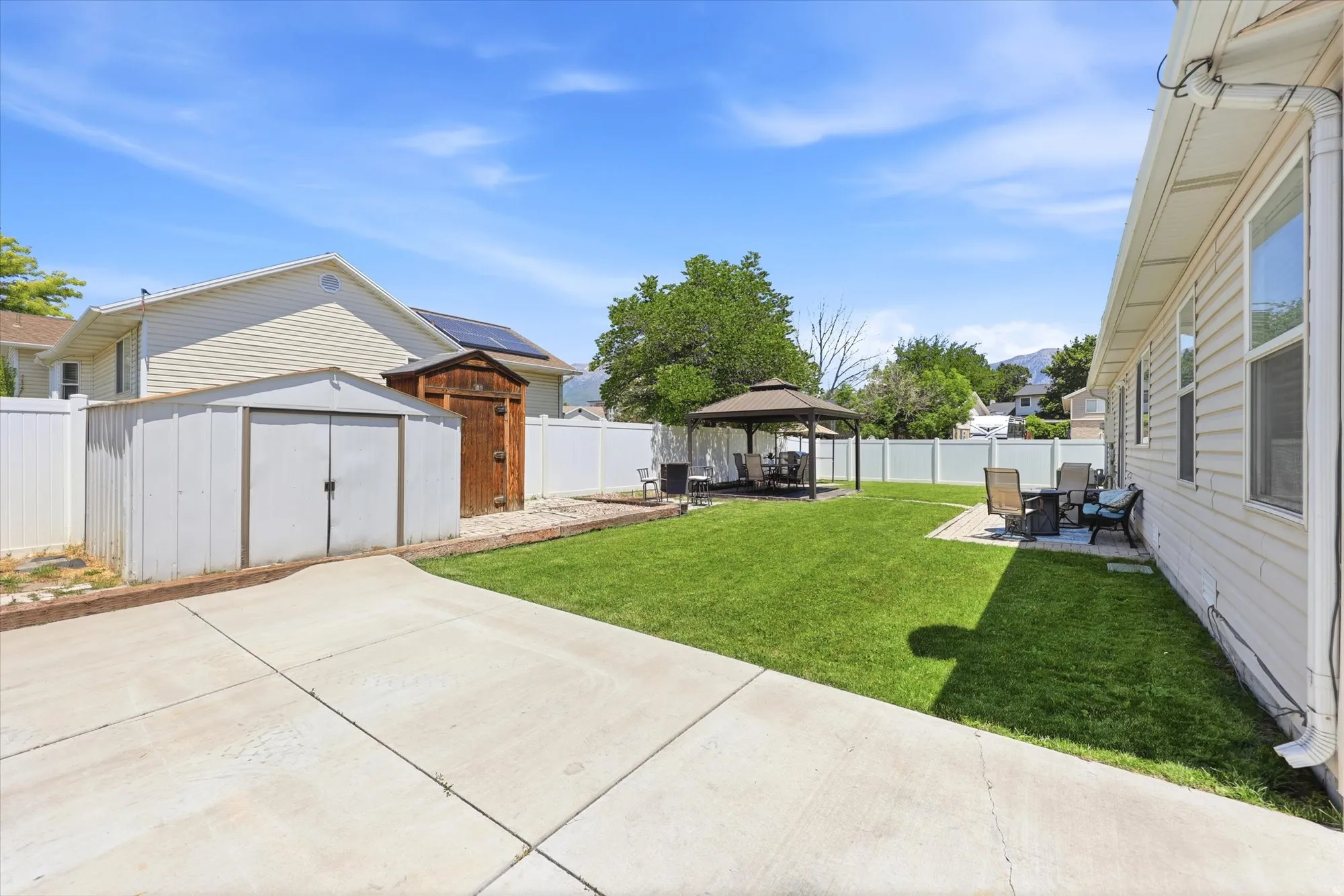 Expansive backyard featuring a large concrete patio, a manicured lawn, a detached gazebo, and two storage sheds