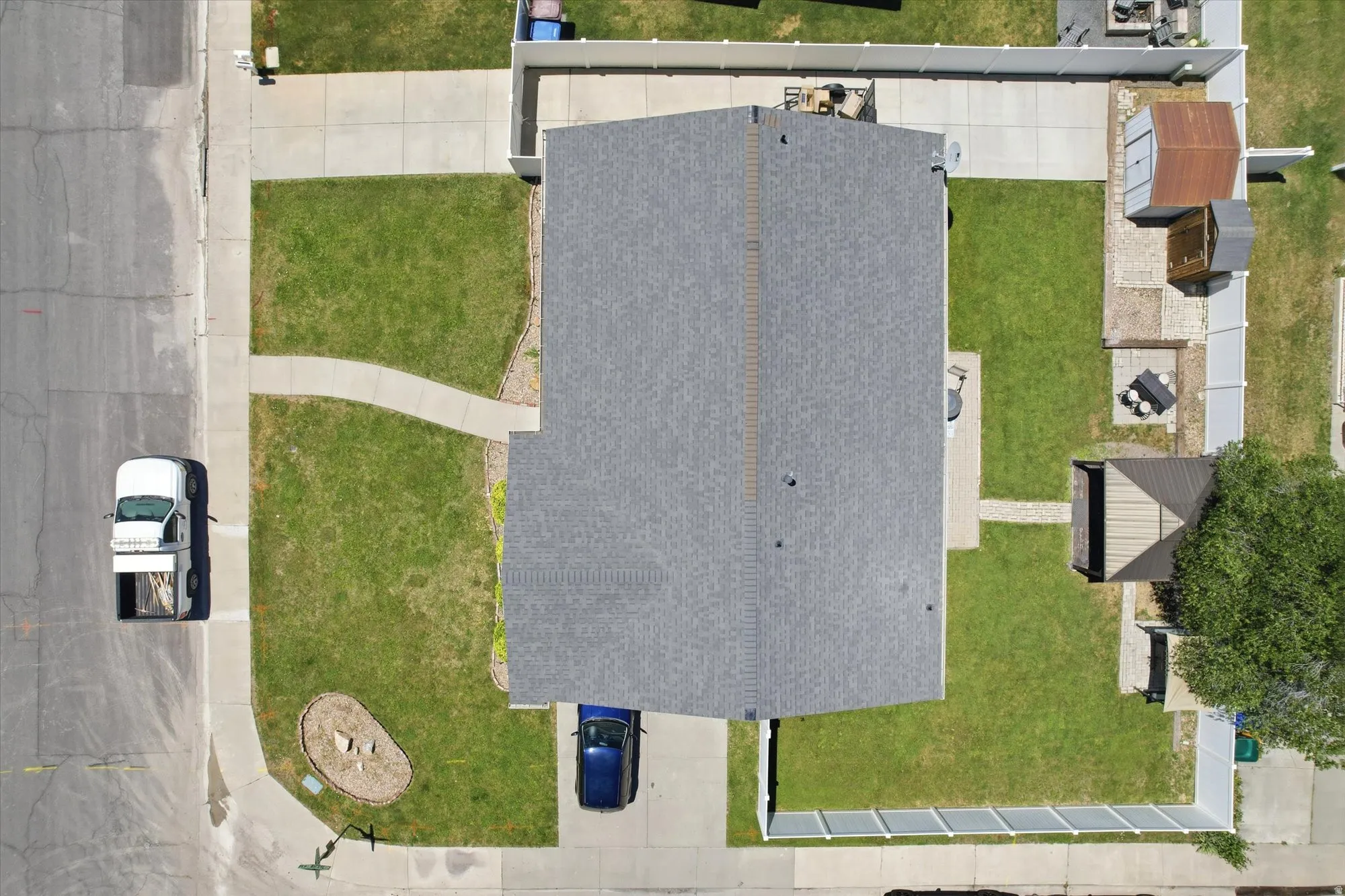 Aerial perspective showcasing the property's layout, featuring a dark gray shingle roof, a concrete driveway, a front lawn with a curved concrete path, and a fenced backyard with a patio