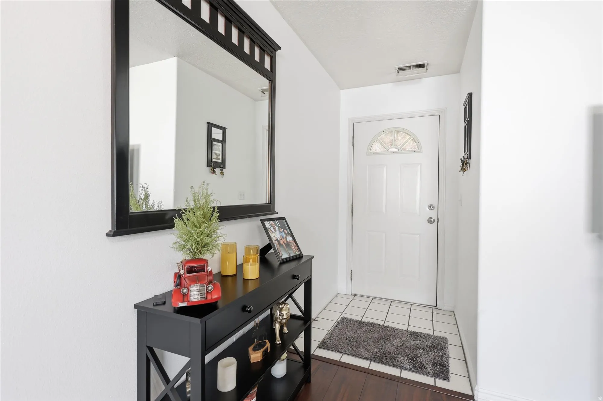 Entryway featuring a white paneled door with an arched decorative glass insert, a black framed mirror, a black console table with drawers and open shelving, white walls, and a combination of tile and wood-finish flooring