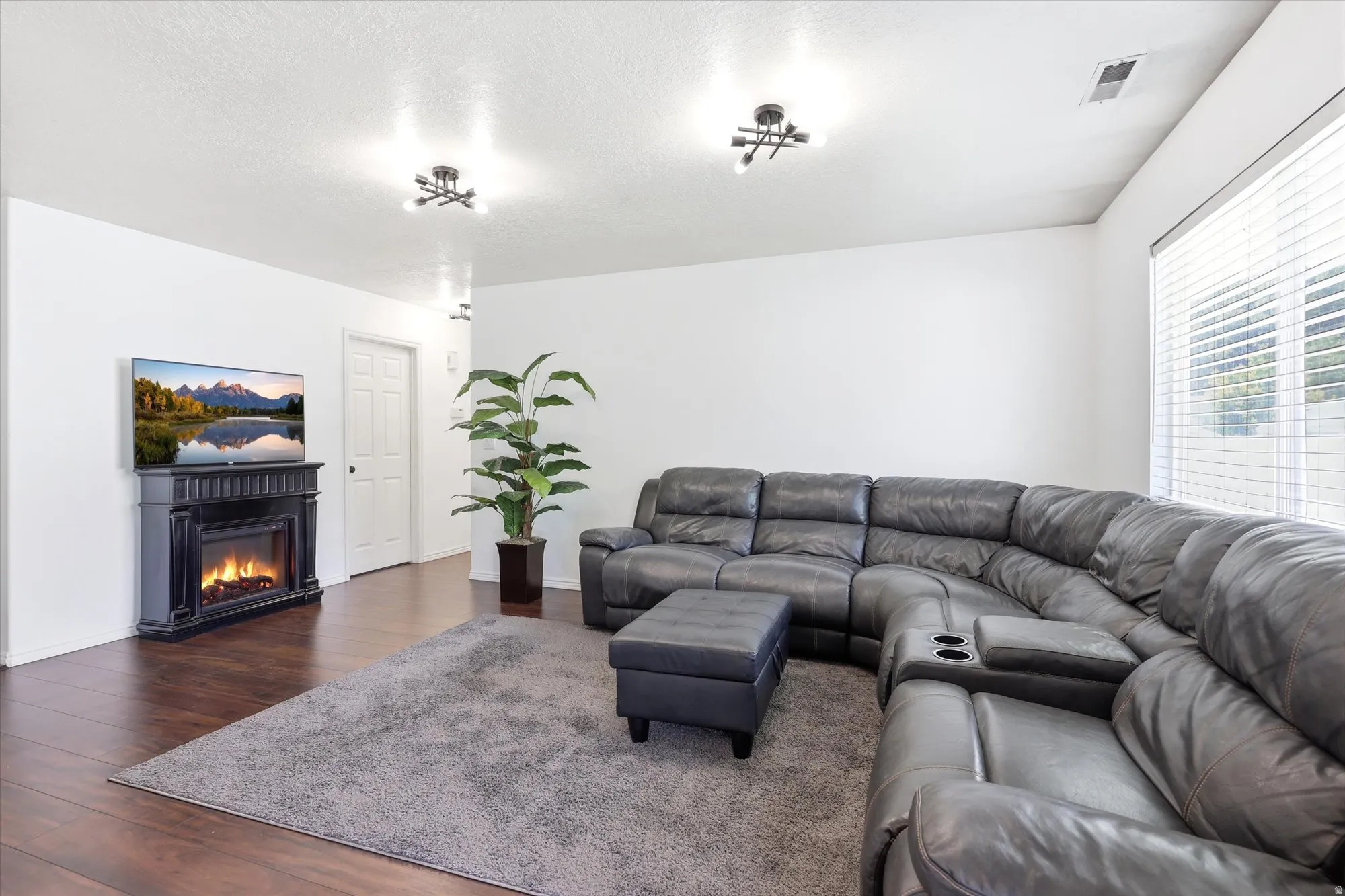 Spacious living area featuring dark wood-finish flooring, a decorative fireplace with mantel, and window blinds