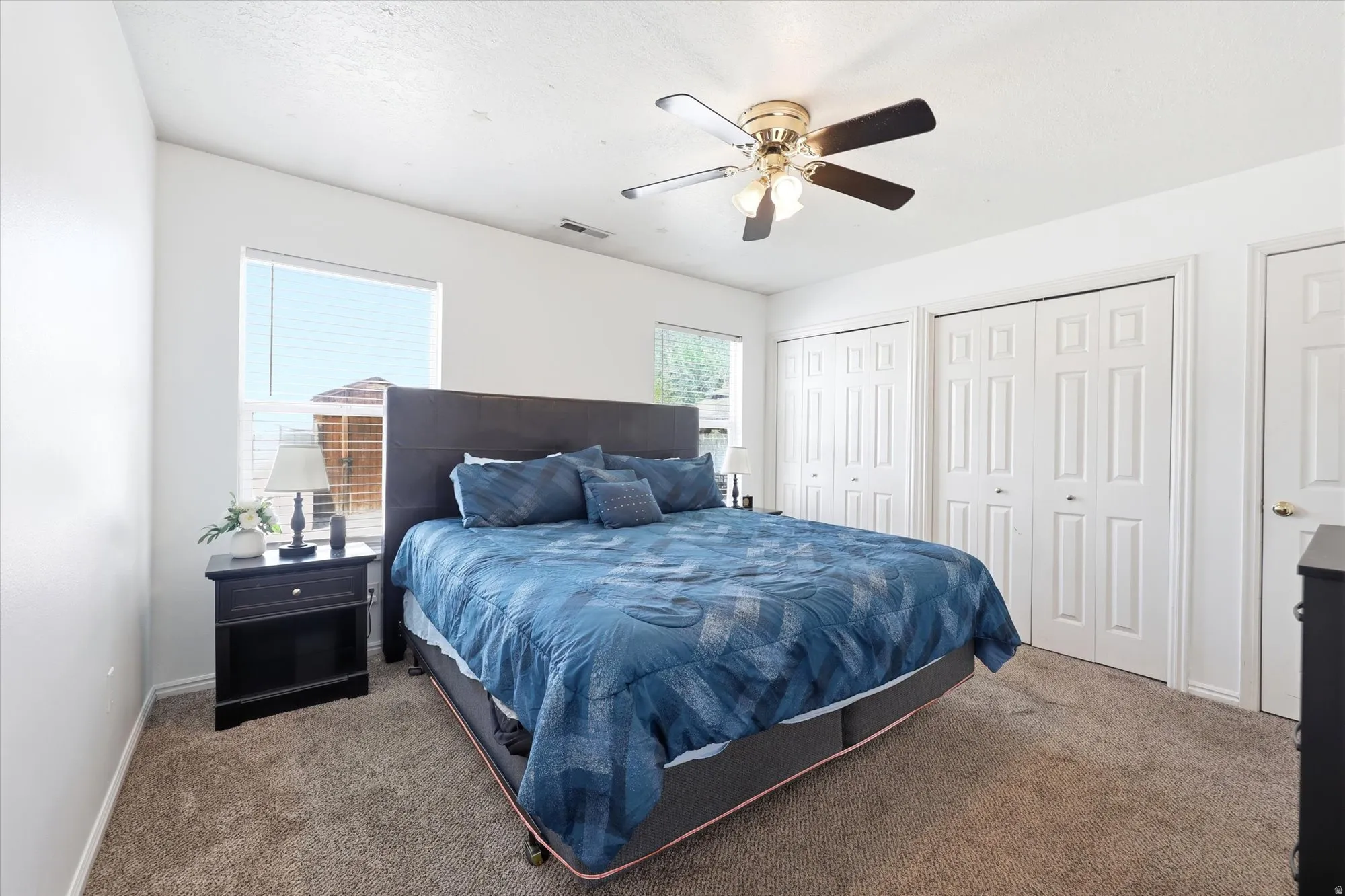 Carpeted bedroom with a ceiling fan light fixture, two windows, and two bi-fold closet doors