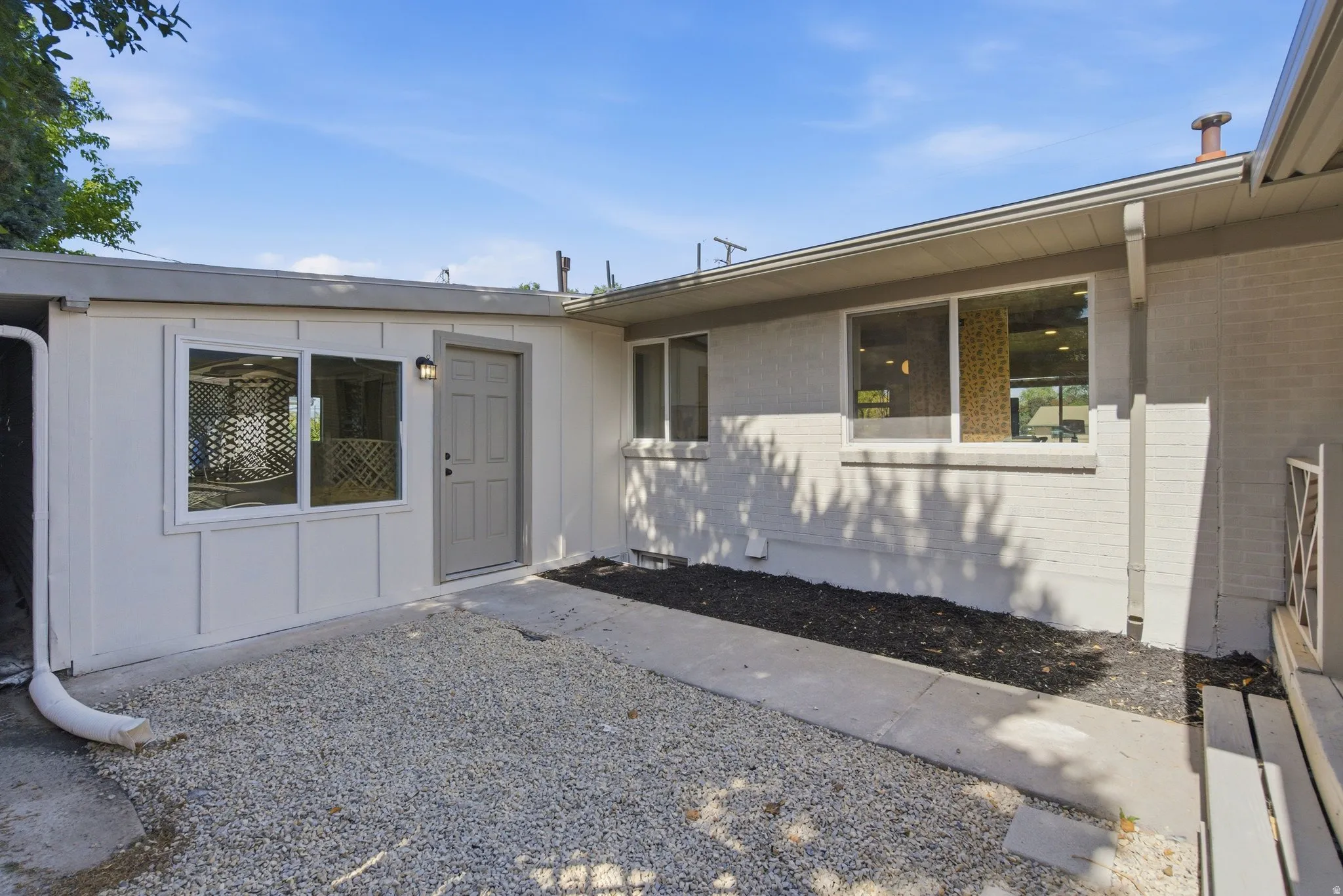Exterior featuring a paneled door with an overhead light fixture, multiple windows, and a gravel-covered area