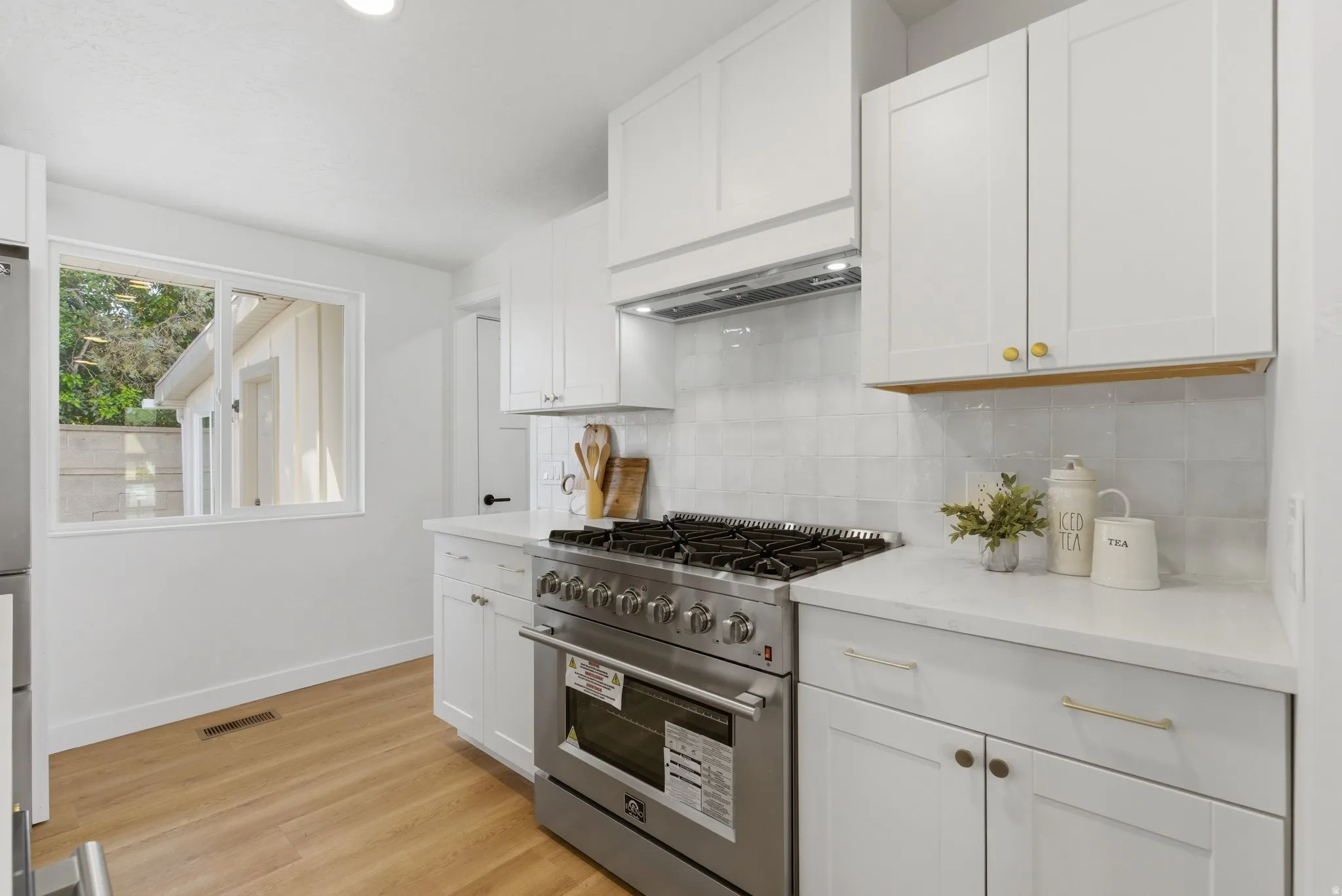 Modern kitchen featuring white shaker cabinetry, stainless steel range with hood, light-toned wood-finish flooring, white countertops, and a subway tile backsplash