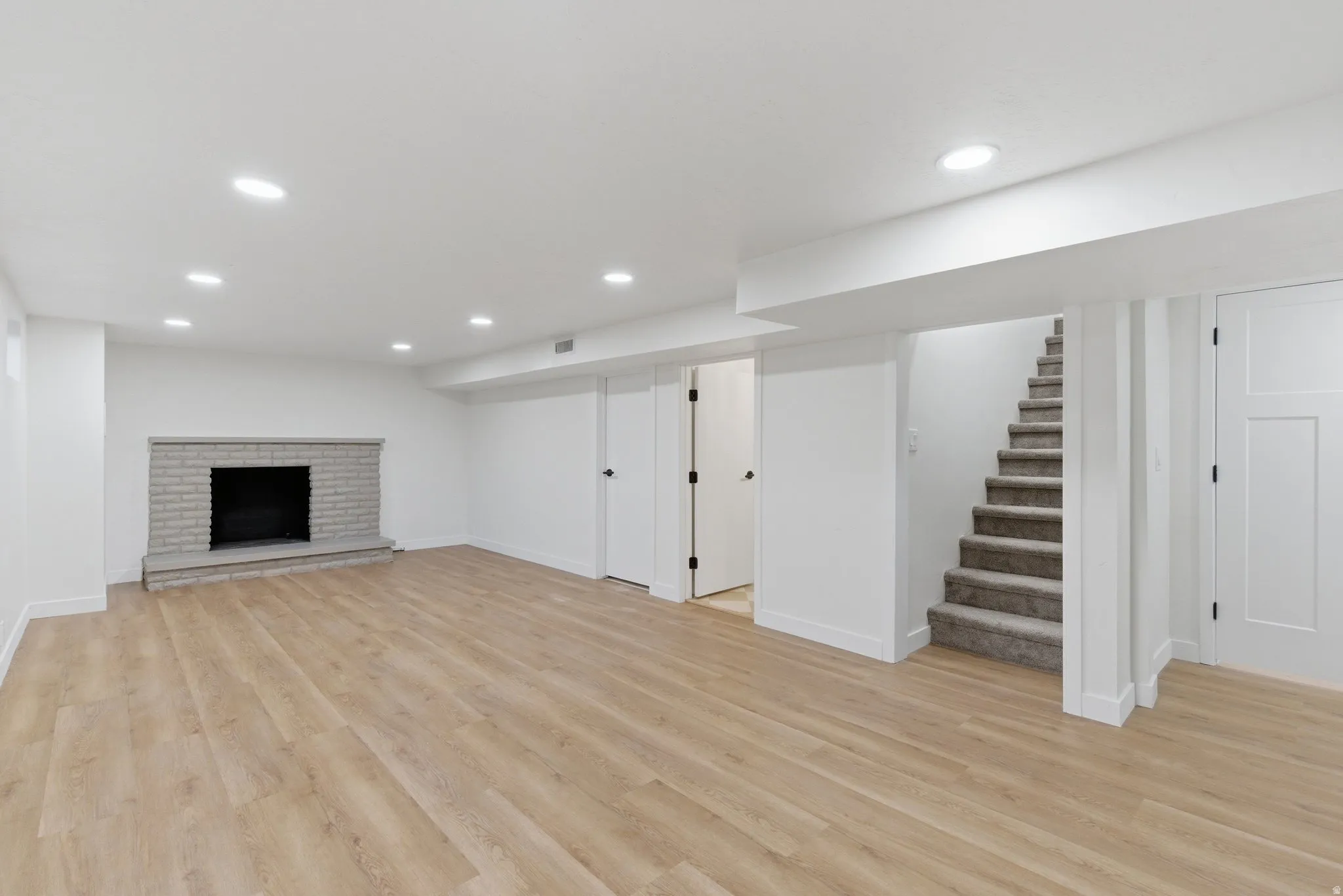 Spacious room featuring wood-finish flooring, a light gray brick fireplace, recessed lighting, and a carpeted staircase