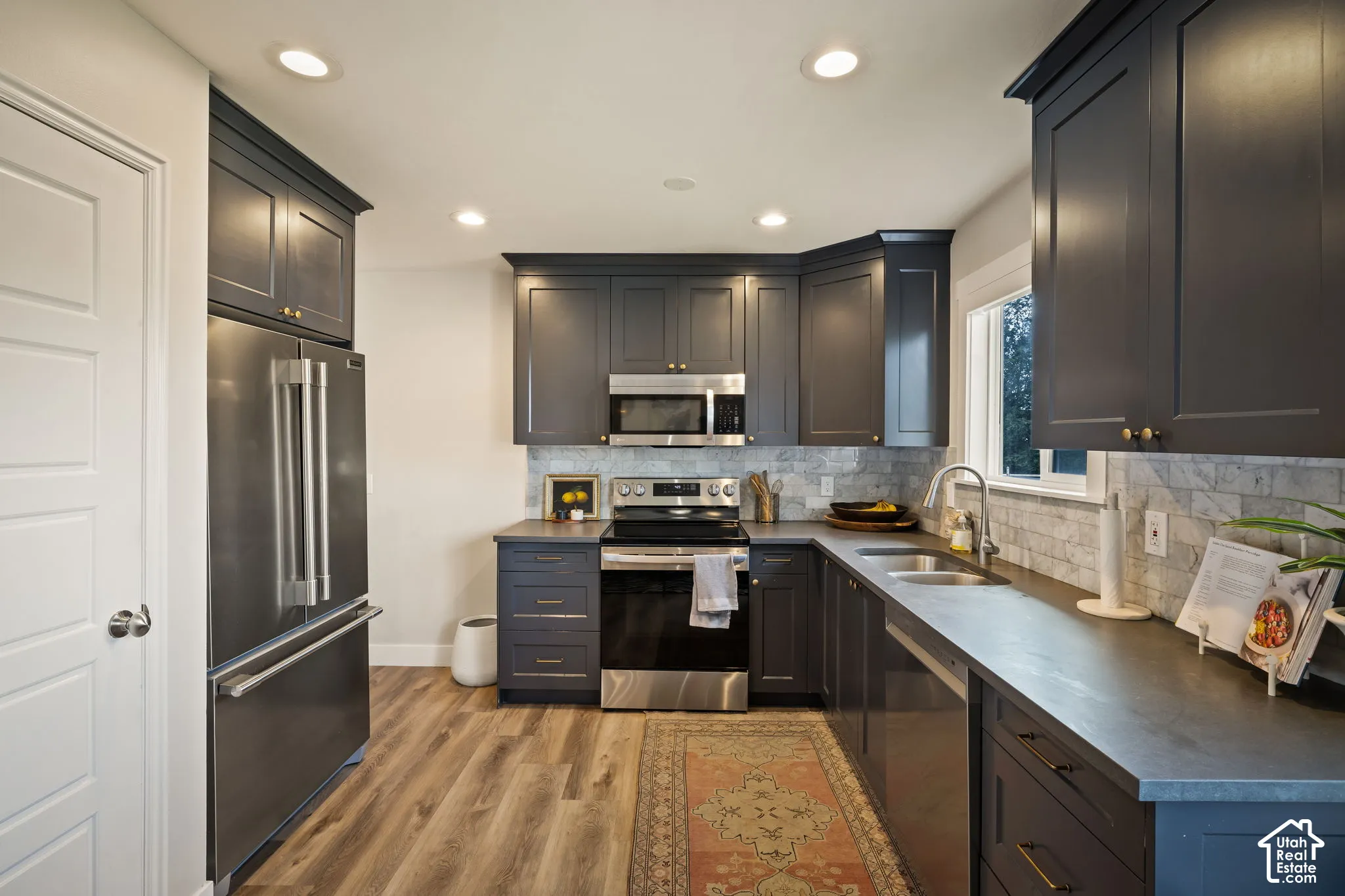 Kitchen featuring dark-toned cabinetry, stainless steel appliances, wood-finish flooring, recessed lighting, and a window above the sink