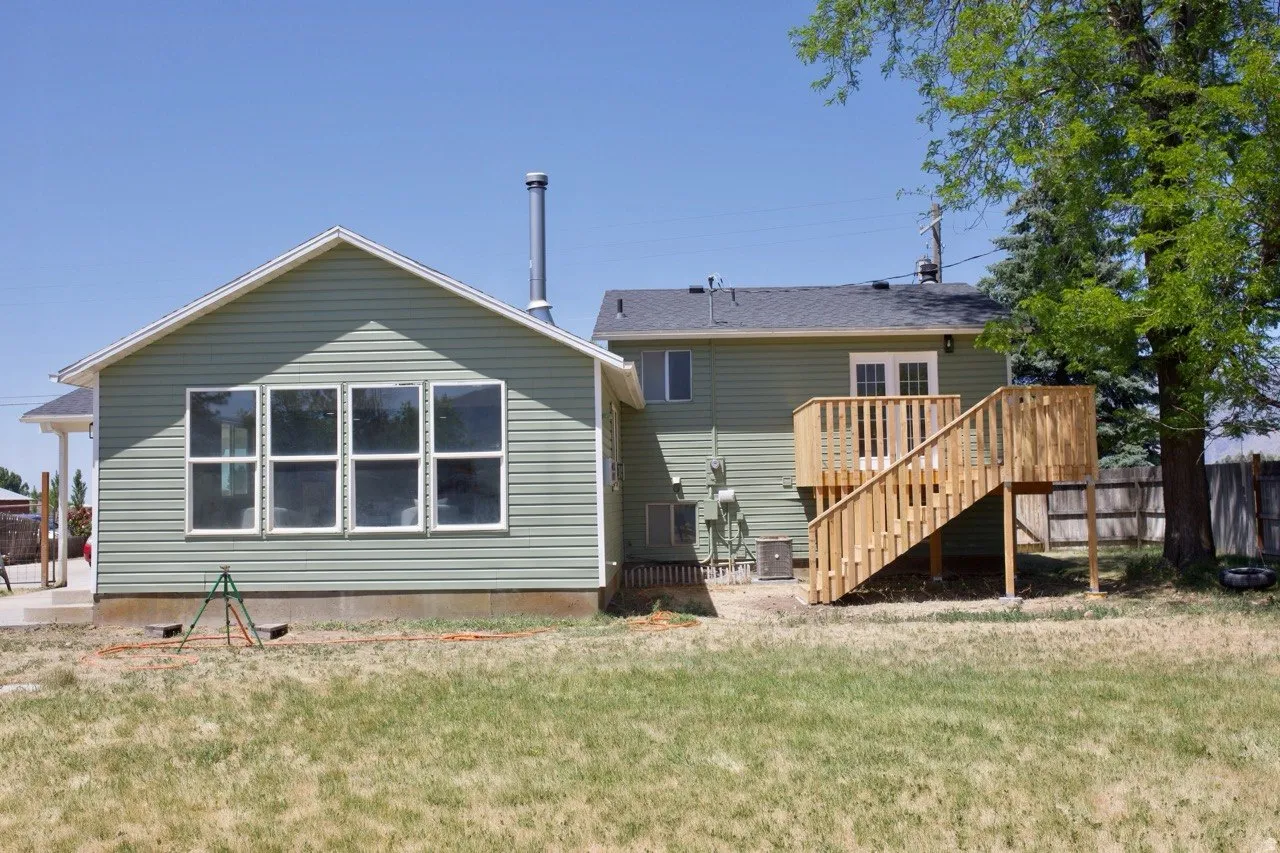 Rear exterior featuring green siding, a gable roofline, multiple pane windows, a wood deck with railings, and a fenced perimeter