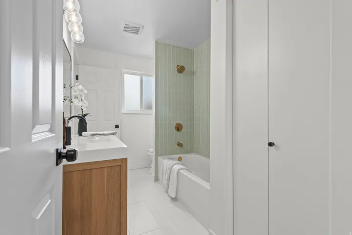 Spa-inspired bathroom featuring a wood-finish vanity with integrated sink and matte black hardware, white subway tile walls, and a soaking tub with green vertical tile surround