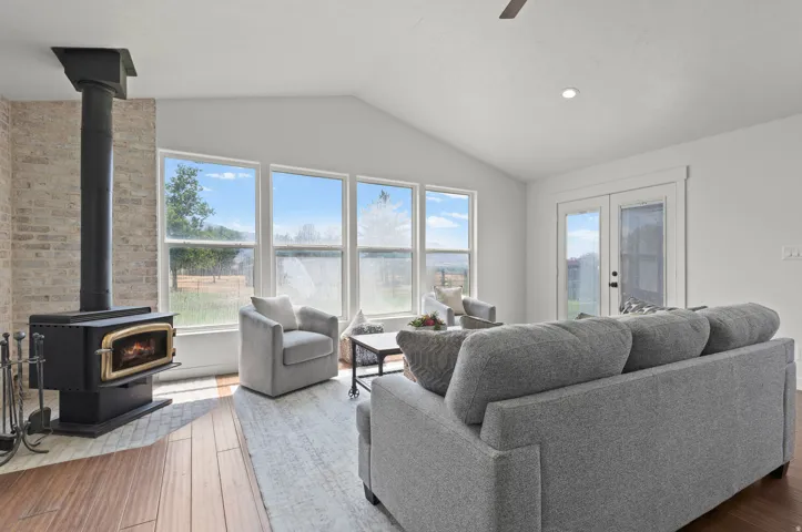 Vaulted ceiling with recessed lighting, wood-burning stove with brick surround, wood-finish flooring, large windows, and double glass-paneled doors