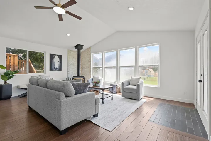Vaulted ceiling with ceiling fan, expansive windows, wood-finish flooring, built-in wood-burning stove, and a stone-clad wall accent