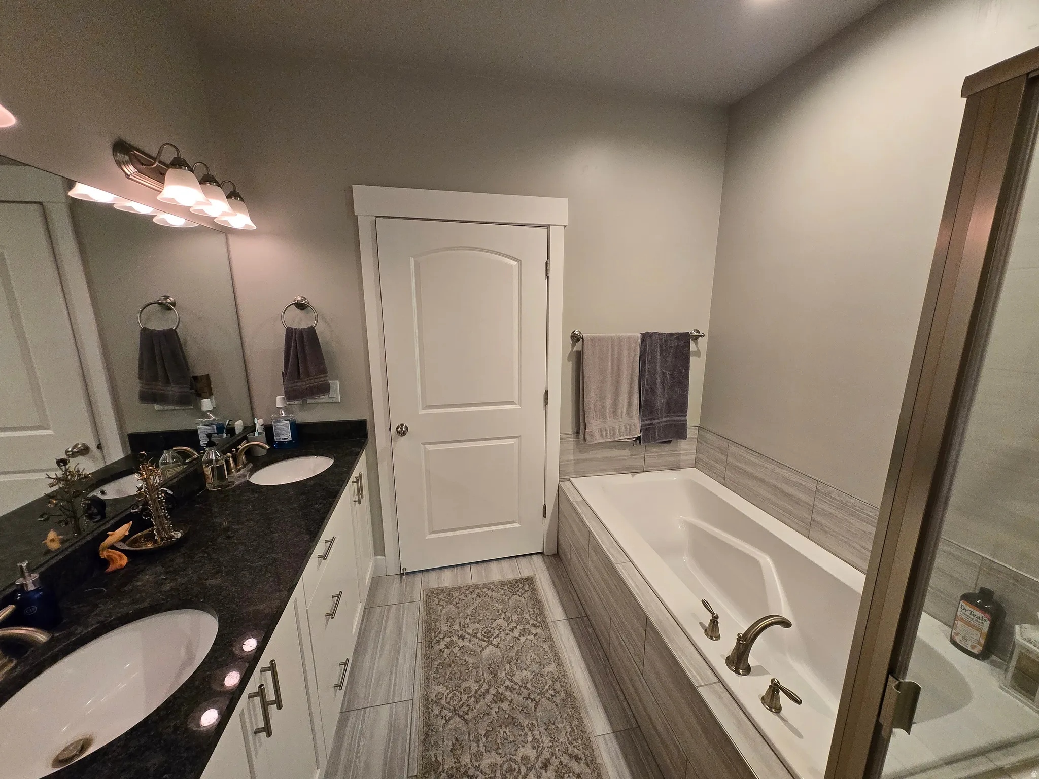 Bathroom featuring a double vanity with dark countertops and dual undermount sinks, a built-in soaking tub with tile surround, and an enclosed shower with a bronze-toned frame