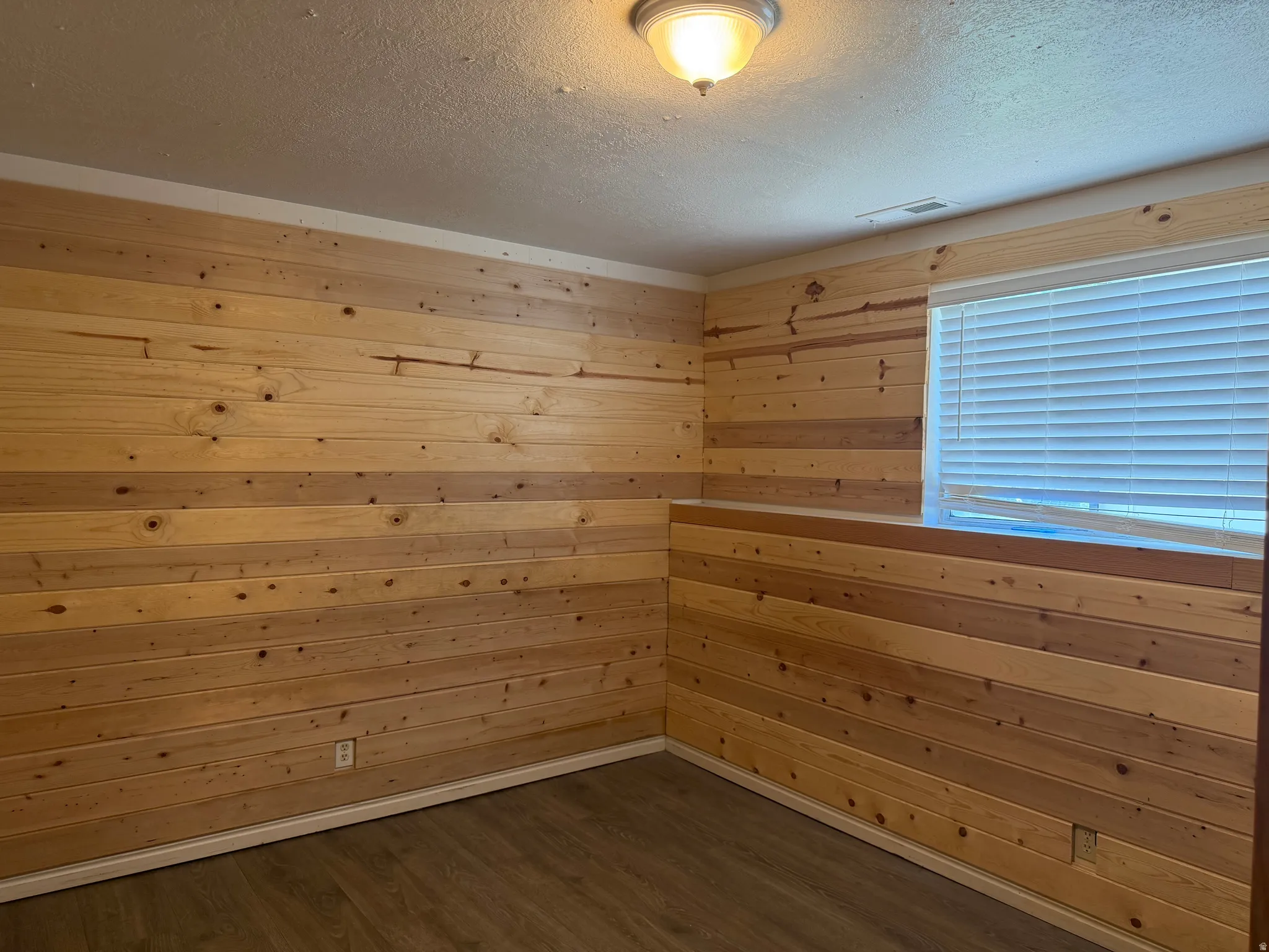 Room featuring horizontal wood paneling, wood-finish flooring, a window with blinds, a white ceiling, and a flush-mount light fixture