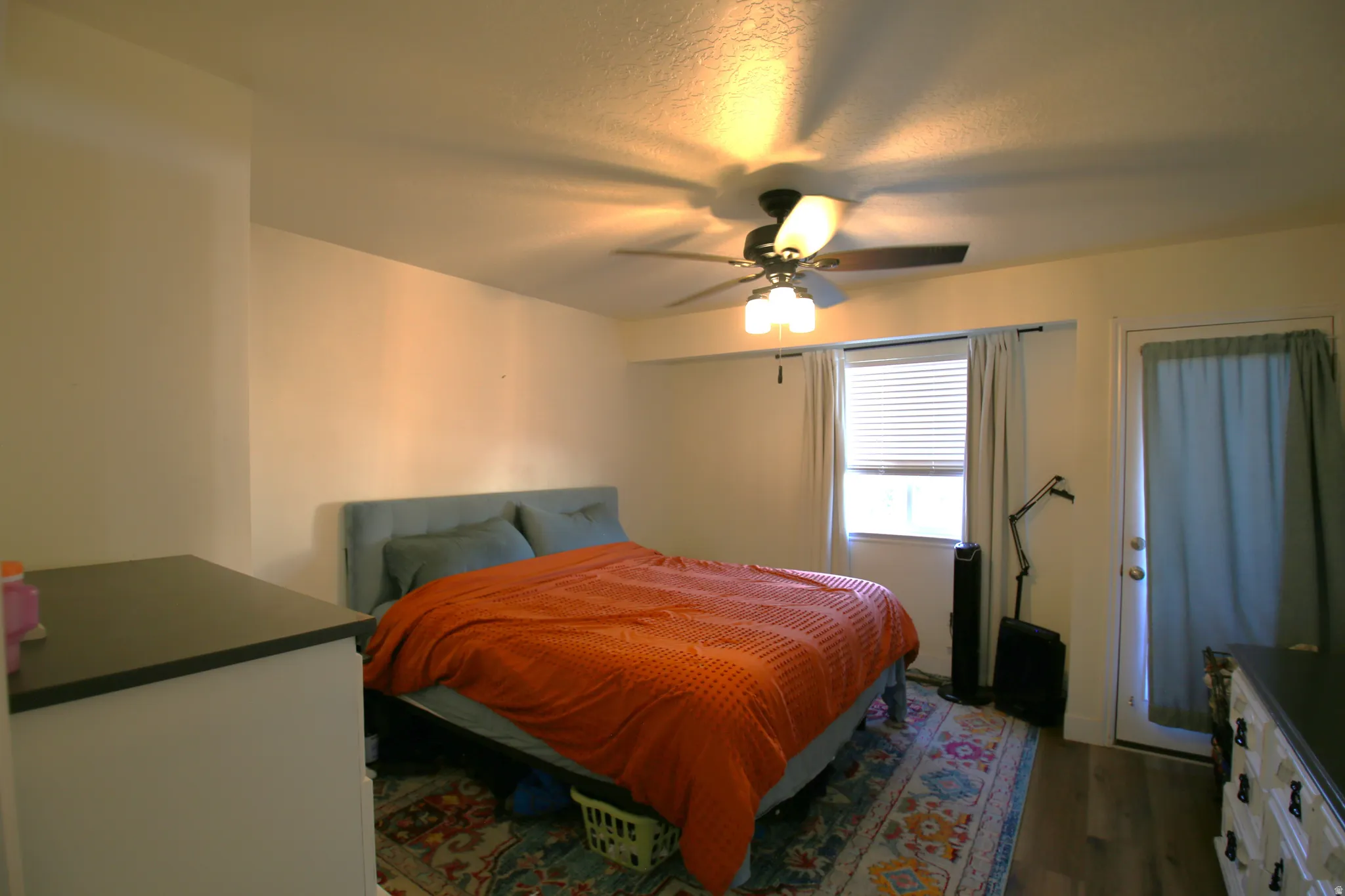 Bedroom featuring a ceiling fan with integrated lighting, wood-finish flooring, a window with blinds, and a door providing exterior access