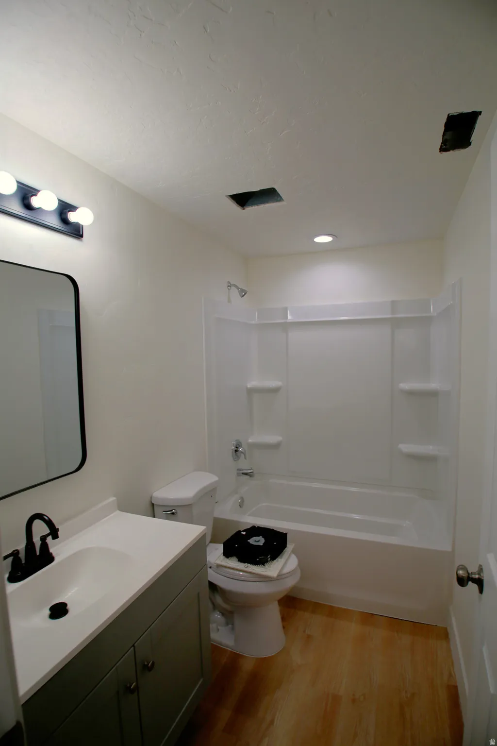 Bathroom featuring light wood-finish flooring, a white vanity with integrated sink, a dark-framed rectangular mirror, a white toilet, and a white bathtub with a shower surround