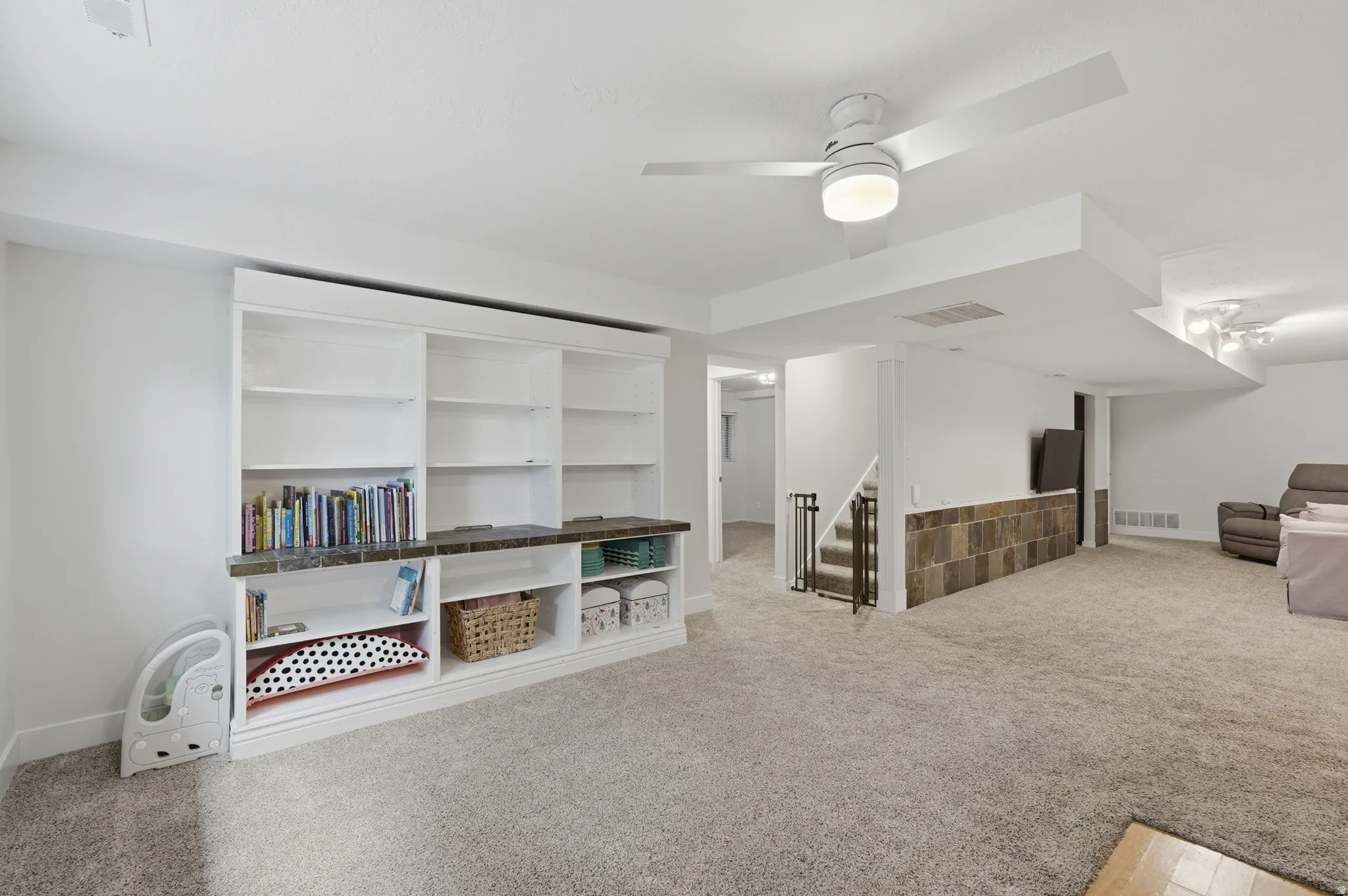 Spacious room featuring extensive built-in shelving, plush carpet flooring, a ceiling fan with integrated lighting, and a partial wall finished with stone-look tile